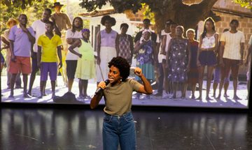 A woman with microphone in front of large photo of people in a line