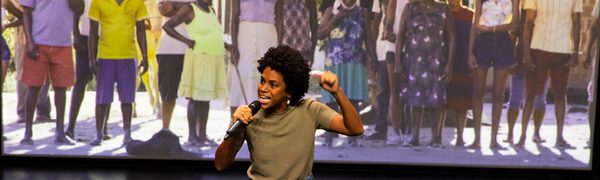 A woman with microphone in front of large photo of people in a line
