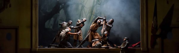 Two men dressed in armour holding sword over another, others reaching inwards towards them in framed scene