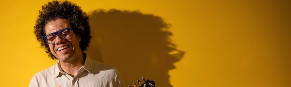 Man laughing holding stringed instrument, standing against yellow wall with bold shadows