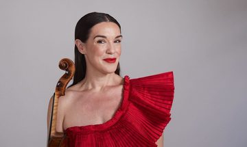 A dark-haired woman wearing a red dress smiles into the camera, a violin at her side