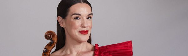 A dark-haired woman wearing a red dress smiles into the camera, a violin at her side