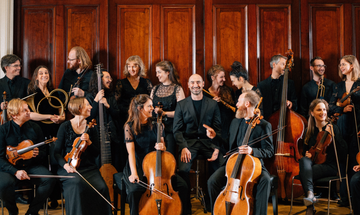 Group of people standing holding stringed instruments, all looking at man centre stage who is facing camera