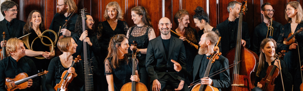 Group of people standing holding stringed instruments, all looking at man centre stage who is facing camera