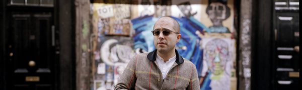 A man wearing dark sunglasses stands in front of a graffitied door holding a trilby in his hand