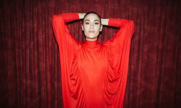 A woman in a draping red dress stands against a deep red curtain, looking into the camera with her hands behind her head
