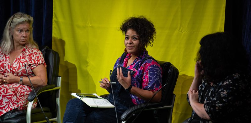 A women sits on a chair with a notebook on her lap. She is talking to a group of people