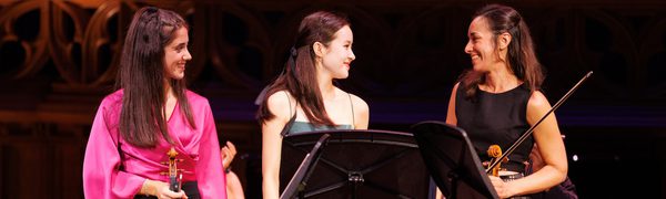 Three woman looking at each other smiling and holding stringed instruments