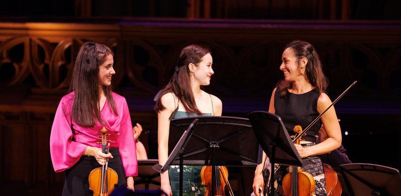 Three woman looking at each other smiling and holding stringed instruments