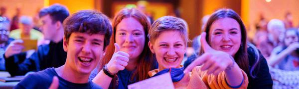 Four teenagers sit in a crowded venue smiling with their thumbs pointed up gesturing towards a ticket