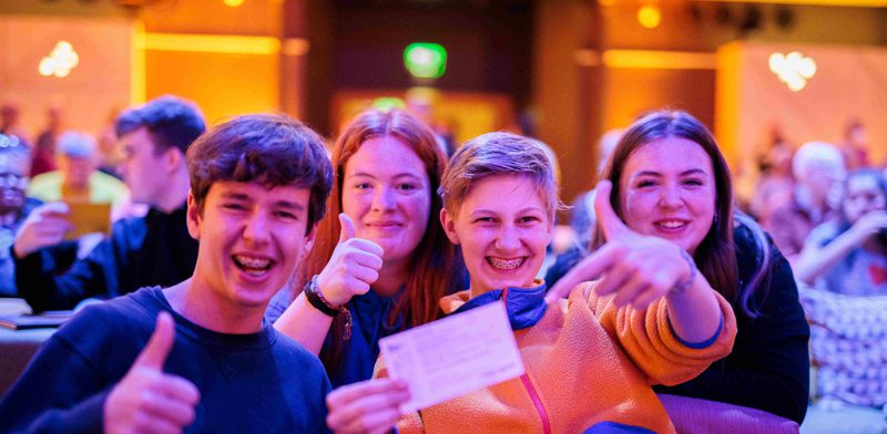 Four teenagers sit in a crowded venue smiling with their thumbs pointed up gesturing towards a ticket