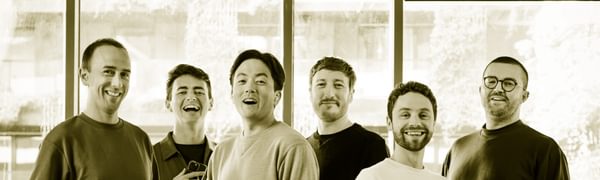 Six singers stand facing the camera dressed in casual clothes