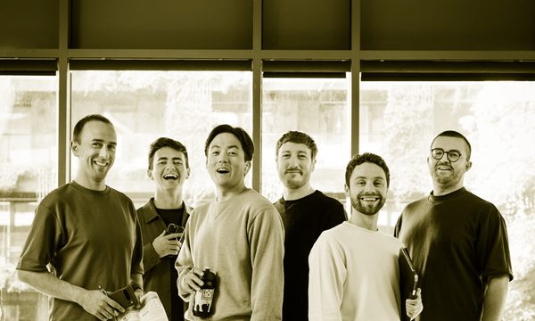 Six singers stand facing the camera dressed in casual clothes