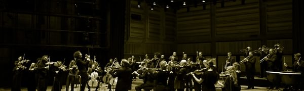 An orchestra dressed in black stands on stage playing their instruments, a conductor stands before them and as an audience looks on