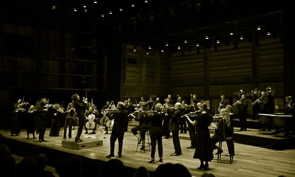 An orchestra dressed in black stands on stage playing their instruments, a conductor stands before them and as an audience looks on