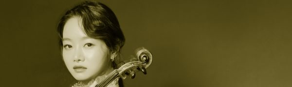 A woman looks at the camera facing side on, she is holding a violin on her shoulder and wearing a pink dress, while standing in front of a blue background