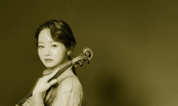 A woman looks at the camera facing side on, she is holding a violin on her shoulder and wearing a pink dress, while standing in front of a blue background