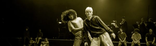 Two dancers are at the front of the stage, one is standing behind the other with their hand on the other dancer's shoulder. Behind them are other dancers seated and a group of musicians