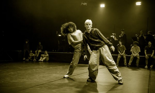 Two dancers are at the front of the stage, one is standing behind the other with their hand on the other dancer's shoulder. Behind them are other dancers seated and a group of musicians