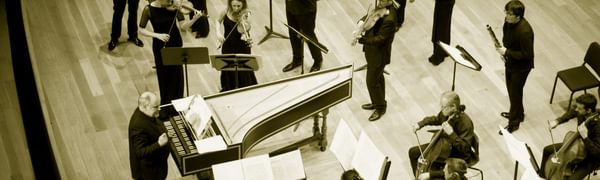 An ensemble of musicians dressed in black stand on stage playing string instruments. The conductor stands in front of them beside a piano.