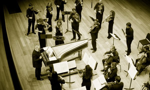 An ensemble of musicians dressed in black stand on stage playing string instruments. The conductor stands in front of them beside a piano.