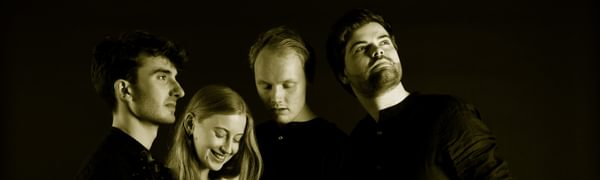 Four musicians stand together all facing different directions. Their black clothing blends into the black background.