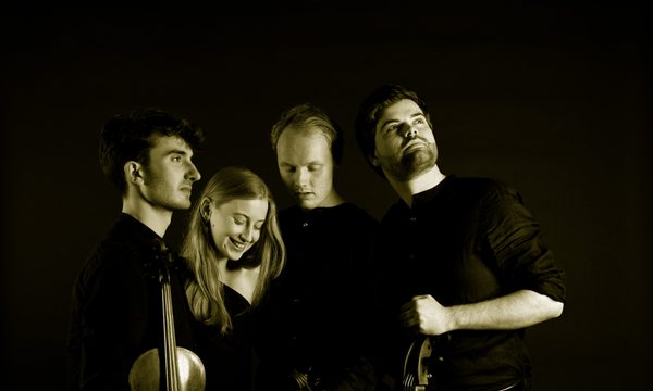 Four musicians stand together all facing different directions. Their black clothing blends into the black background.