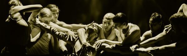 A dancer kneels on the ground with his arms stretched out to the sides. A group of dancers surrounds him reaching out to grab his head, arms and chest.