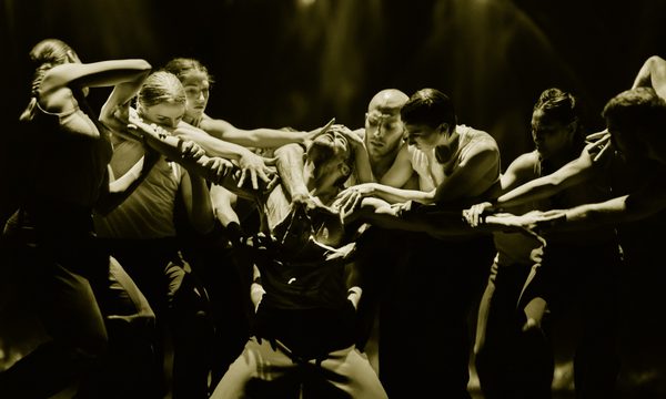 A dancer kneels on the ground with his arms stretched out to the sides. A group of dancers surrounds him reaching out to grab his head, arms and chest.