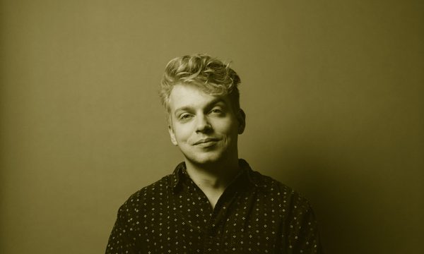 A man sitting facing the camera with one leg crossed over the other. He wears a black spotted shirt and jeans in front of a green background.