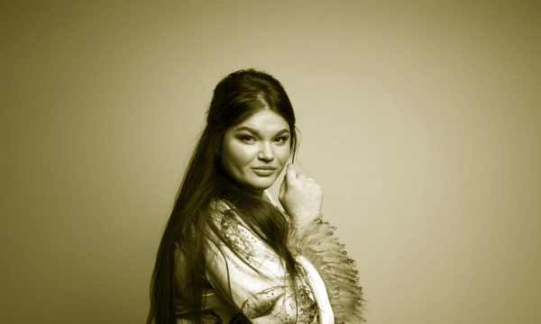 A woman with long brown hair in a pink and white gown looks over her shoulder with a knowing smile