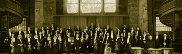 The orchestra stand posed with their instruments in a large room with big windows.