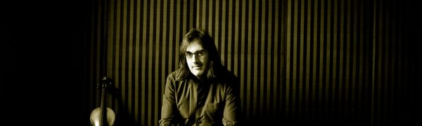 A man with long hair and glasses sits with his hands clasped looking at the viewer. A violin is propped up next to him.