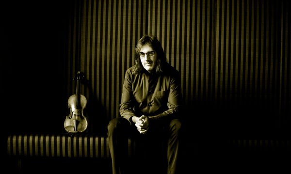 A man with long hair and glasses sits with his hands clasped looking at the viewer. A violin is propped up next to him.