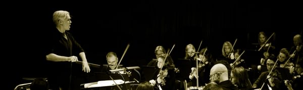 A black and white image of an orchestra sitting on stage. The conductor stands before them pointing at the orchestra.