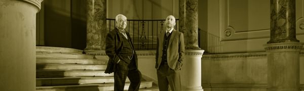 Two men in suits stand looking at the viewer, situated in a grand hall with marble columns