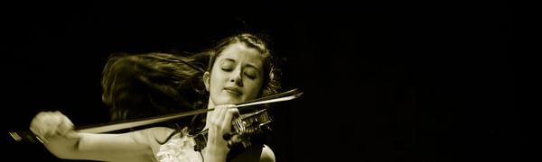A woman dressed in a white dress playing the violin. Her brown hair is flowing behind her.