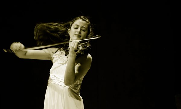 A woman dressed in a white dress playing the violin. Her brown hair is flowing behind her.