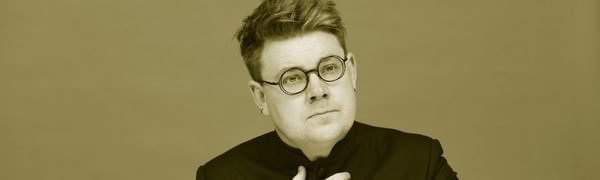 A man with round glasses with both hands held in front of his chest, in black and white