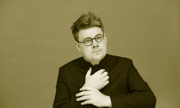A man with round glasses with both hands held in front of his chest, in black and white