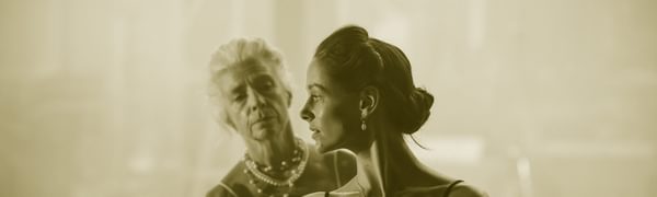 A young woman in black looks over her shoulder. Behind her an older woman in white reaches for her.