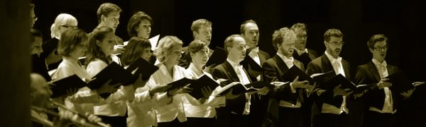 The choir of an orchestra sings, string and brass players visible at the edge of frame.