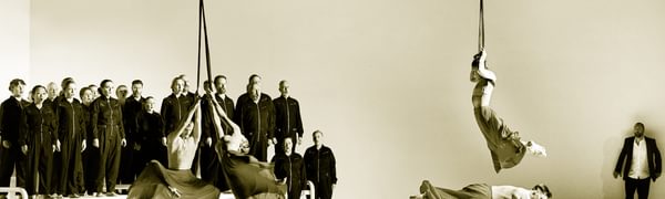 Four dancers perform in red flowing skirts, three of which are suspended from ropes. In the background, a choir dressed in black jumpsuits sing on top of white platforms.