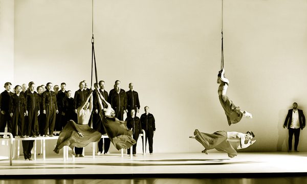 Four dancers perform in red flowing skirts, three of which are suspended from ropes. In the background, a choir dressed in black jumpsuits sing on top of white platforms.