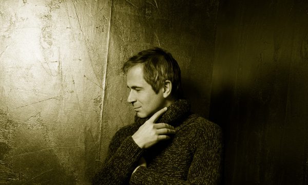 A man stands in a black room facing the side. He is wearing a knitted jumper and black trousers.