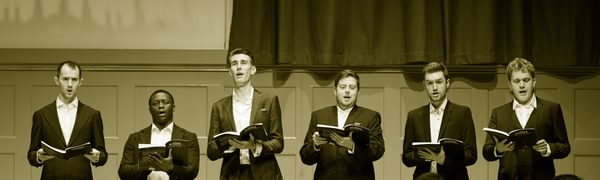 Six men in suits sing on stage holding songbooks.