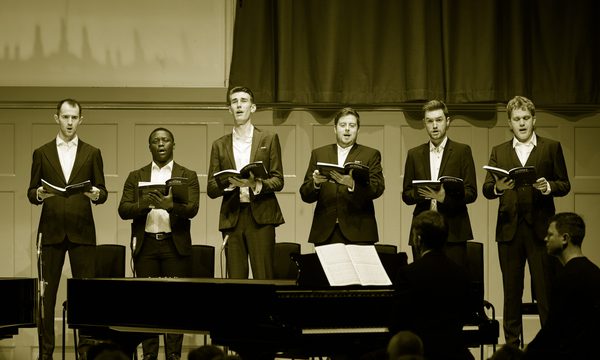 Six men in suits sing on stage holding songbooks.