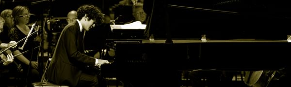 A young man with black hair is playing a grand piano in front of an orchestra