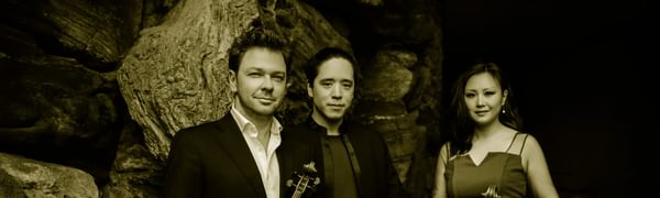 Two men and a woman stand in front of a large rock. The men wear black suits and the woman wears a bright blue dress. They are holding a violin and a cello.