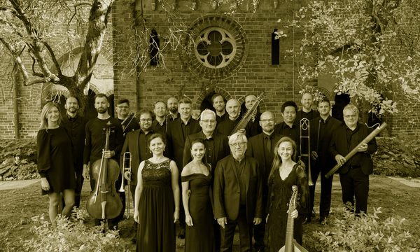 A group of musicians dressed in black standing in front of a brick building. Trees and bushes frame them.